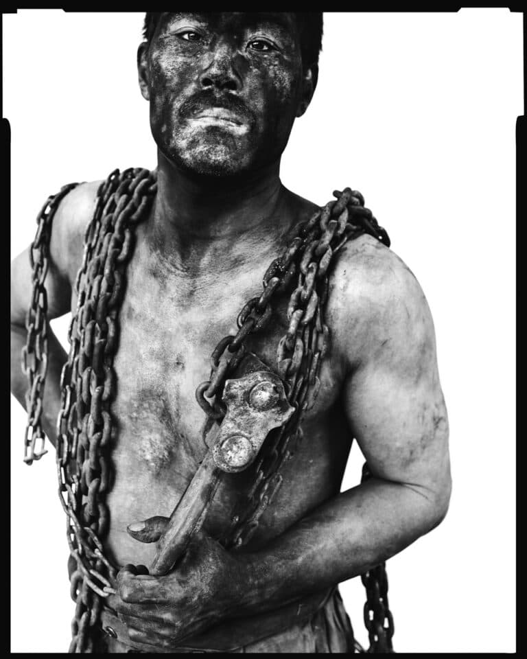Black and white photo of a coal miner covered in coal dust with chains around his neck.