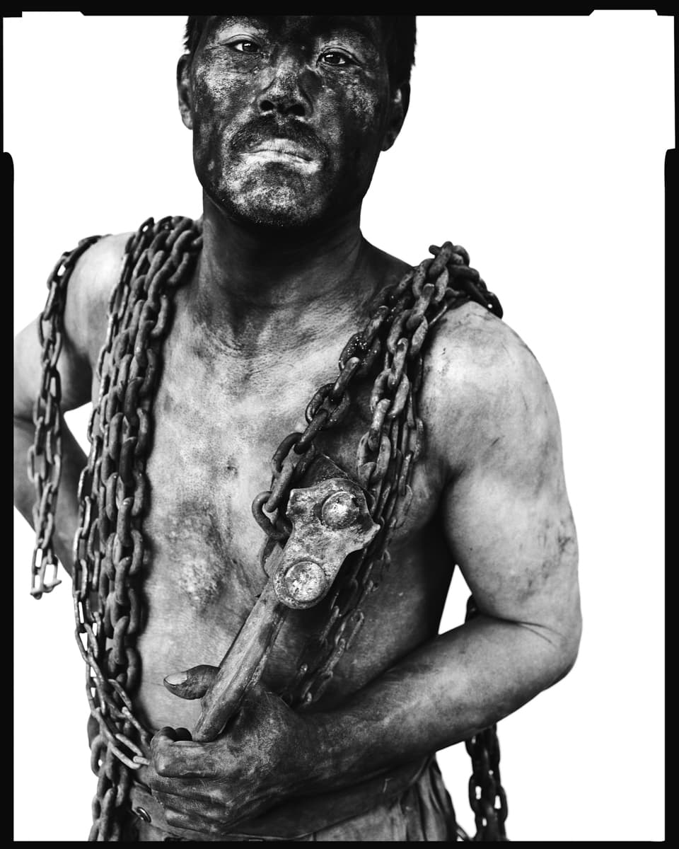 Black and white photo of a coal miner covered in coal dust with chains around his neck.