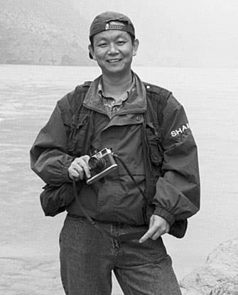 3/4 length B&W portrait of Geng Yunsheng standing by a body of water, wearing a backwards baseball cap, a jacket and vest, and holding a camera in his right hand