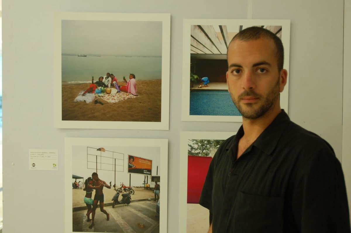 Color photo of Alfredo D'Amato in front of his photographs on a white gallery wall wearing a black button down shirt
