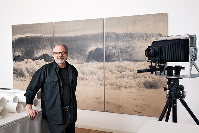 Color photo of Clifford Ross in his studio standing in front of a triptych of his hurricane wave photos, next to a large format camera on a tripod