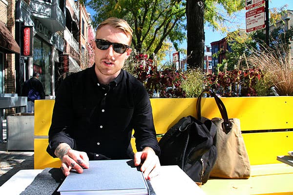 Color photo of Daniel Shea seated on a yellow bench with a grey book in front of him, wearing a black button down shirt and sunglasses