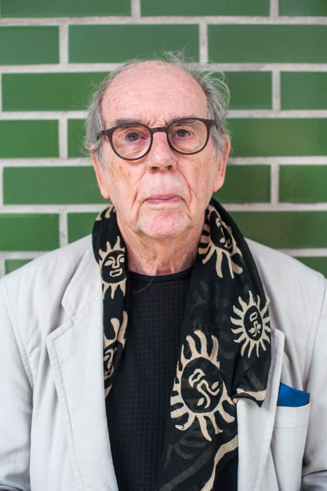 Color quarter length portrait of David Hurn standing in front of a green subway tiled wall wearing a patterned scarf and a light blazer