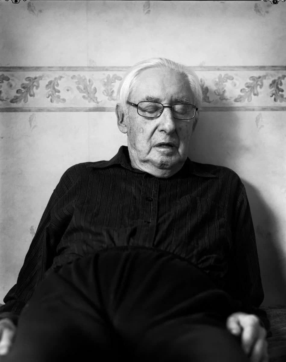 B&W portrait of Dolf Kruger leaning against a wall with eyes closed, wearing wire framed glasses