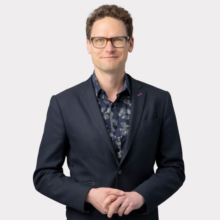 Color portrait of Jake Barton wearing dark framed glasses and a black blazer