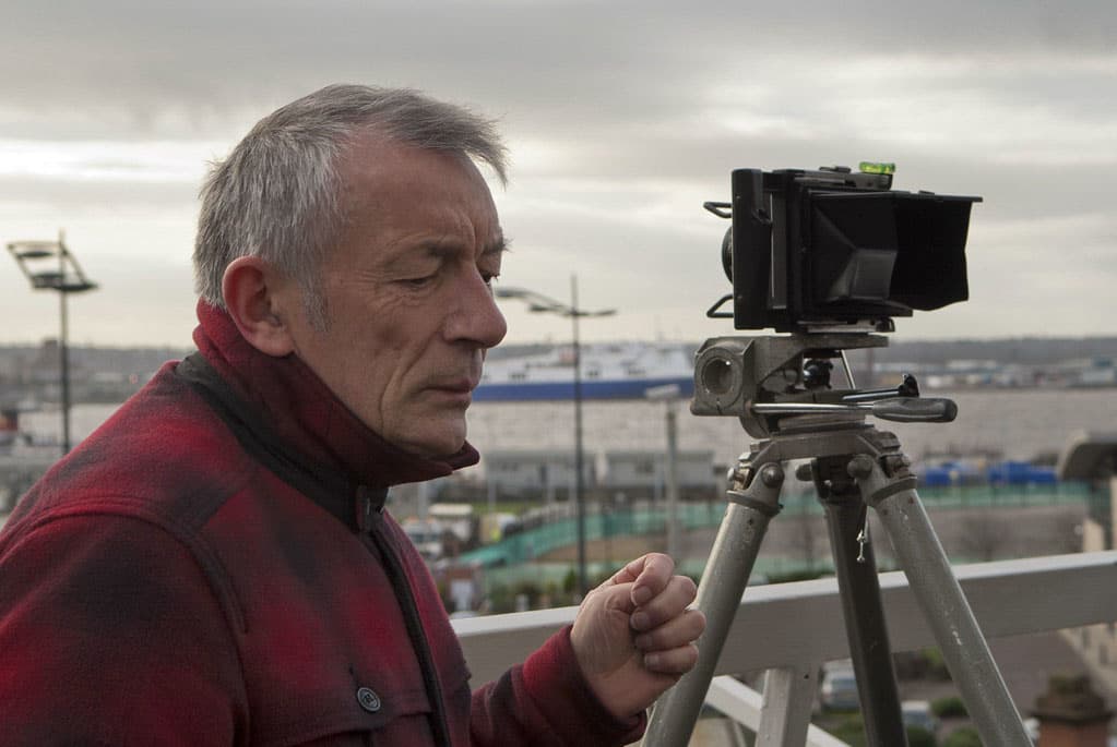 Color photo of John Davies loooking to the right next to a camera on a tripod wearing a flannel jacket