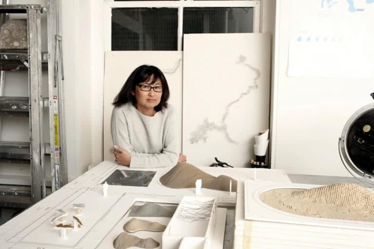 Color photo of Maya Lin, wearing black glasses and a white sweater, seated at a table in her studio, surrounded by architectural models and a ladder