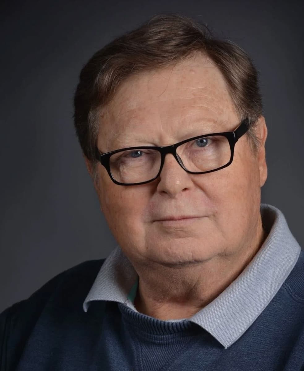 Color portrait of Robert Wallis in front of a dark grey background wearing black framed glasses and a blue collared shirt