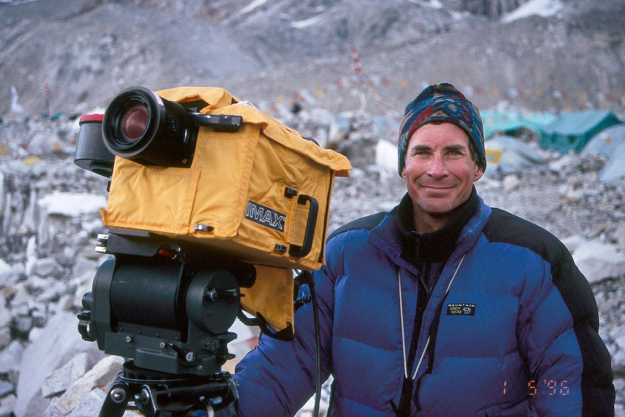 Color photo of David Breashears filming his “Everest” documentary with an IMAX camera, wearing a patterned hat and blue and black puffy coat