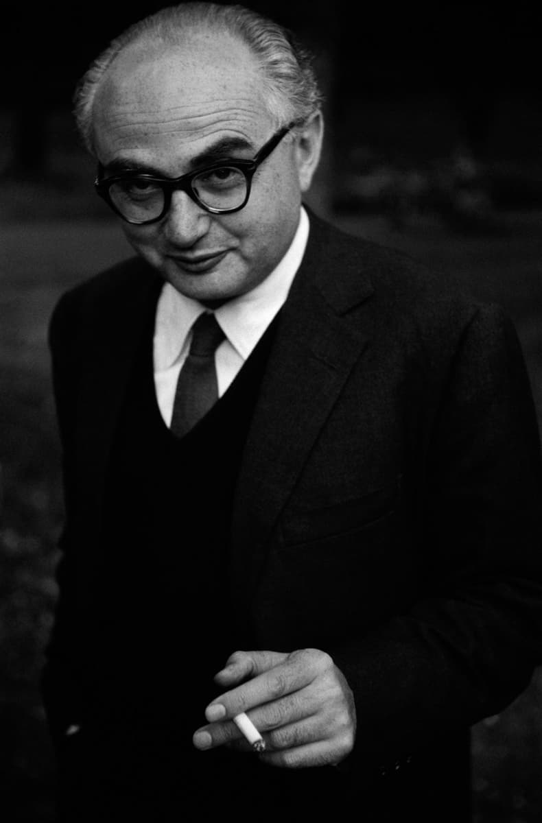B&W half-length portrait of David Seymour wearing dark framed glasses, a suit jacket and tie, and holding a lit cigarette in his left hand