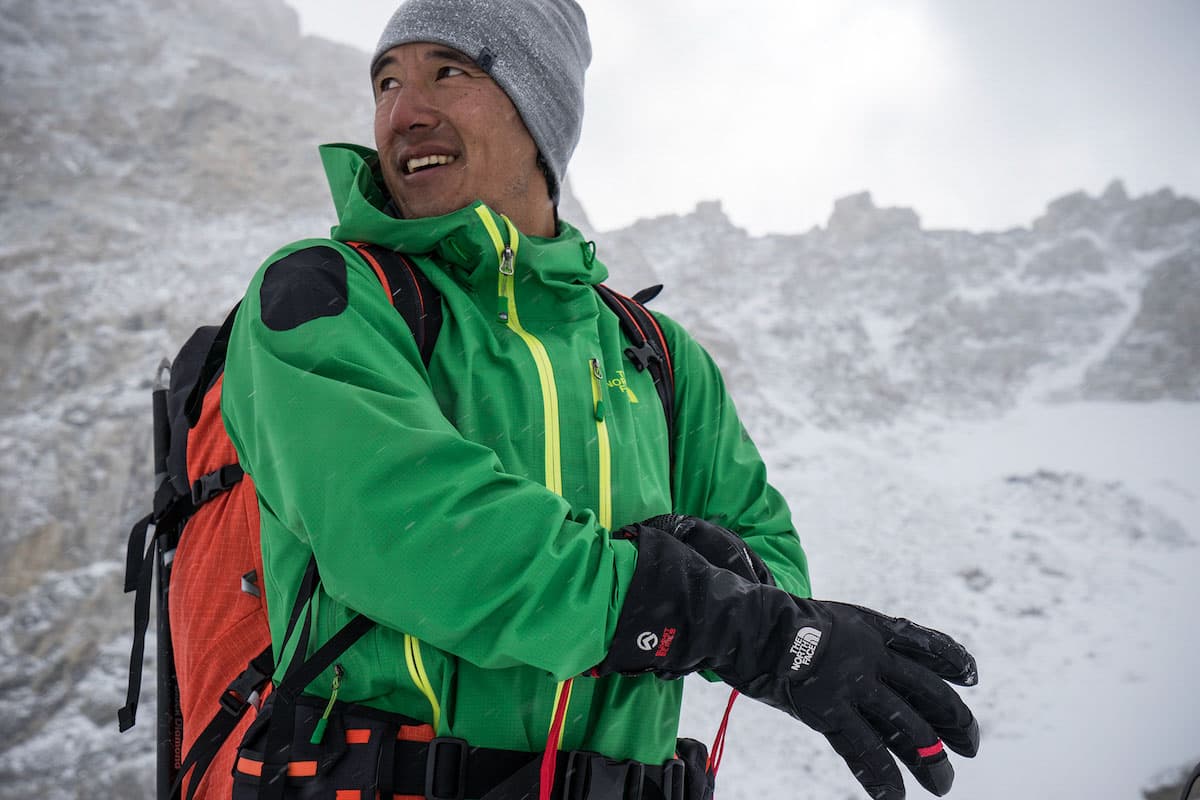 Color photo of Jimmy Chin in snowy mountainous surroundings, looking to the left wearing a grey hat, green jacket, orange pack, and black gloves