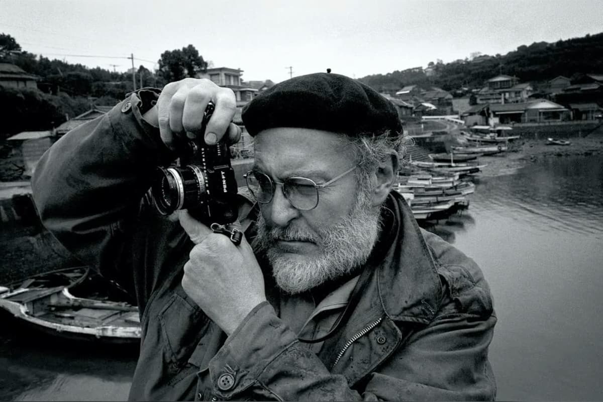 B&W photo of W Eugene Smith in an outdoor setting with boats in the background, hearing a buret, wireframe glasses, and holding a camera up to his right eye