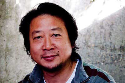 Color portrait of Yu Haibo looking into the camera in front of a stone background wearing a blue zipped shirt