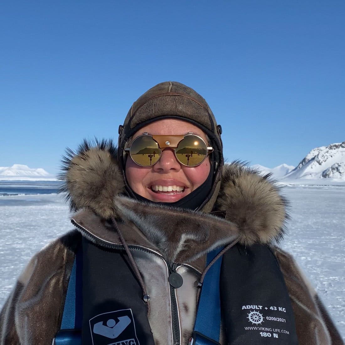 Color self portrait of Camille Seaman in the snow, wearing reflective snow goggles, a leather hat, and a coat with a fur vest