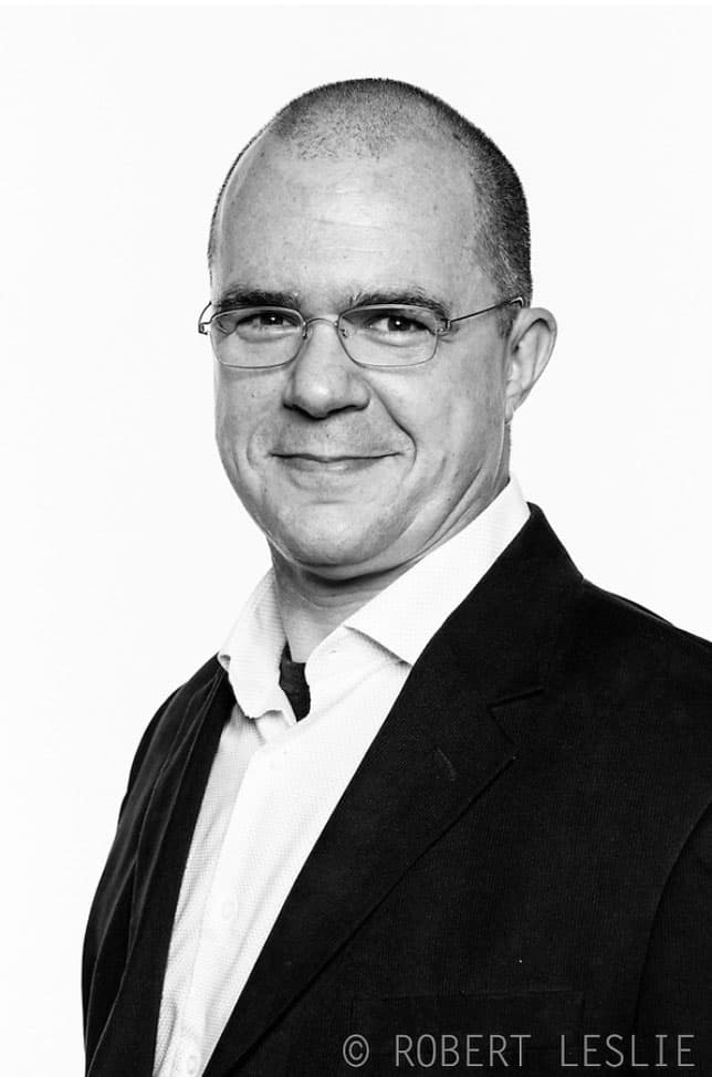 B&W portrait of Daniel Beltra wearing wire framed glasses, a white button down shirt and a dark blazer