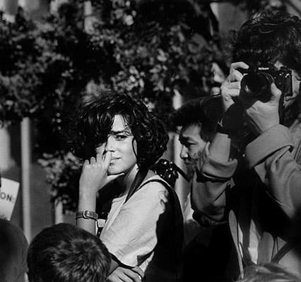 B&W photo of Darcy Padilla, standing among a crowd of people, looking at the camera and holding her chin with her left hand
