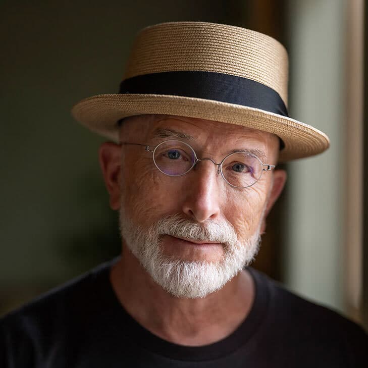 Color portrait of Jamey Stillings wearing wire rimmed glasses, a straw hat, and a black shirt