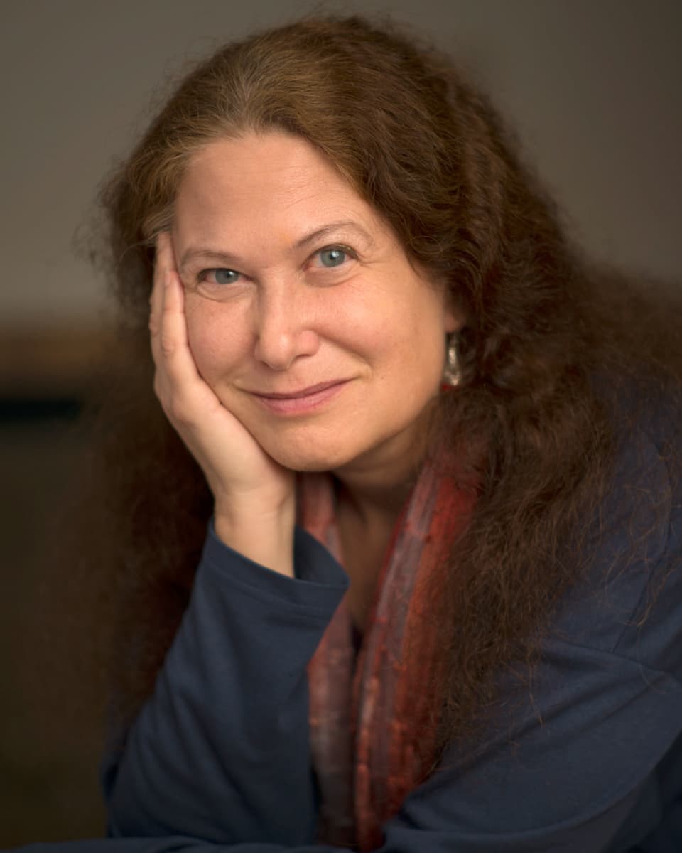 Color portrait of Jane Hirshfield wearing a blue long sleeve shirt and holding her face with her right hand
