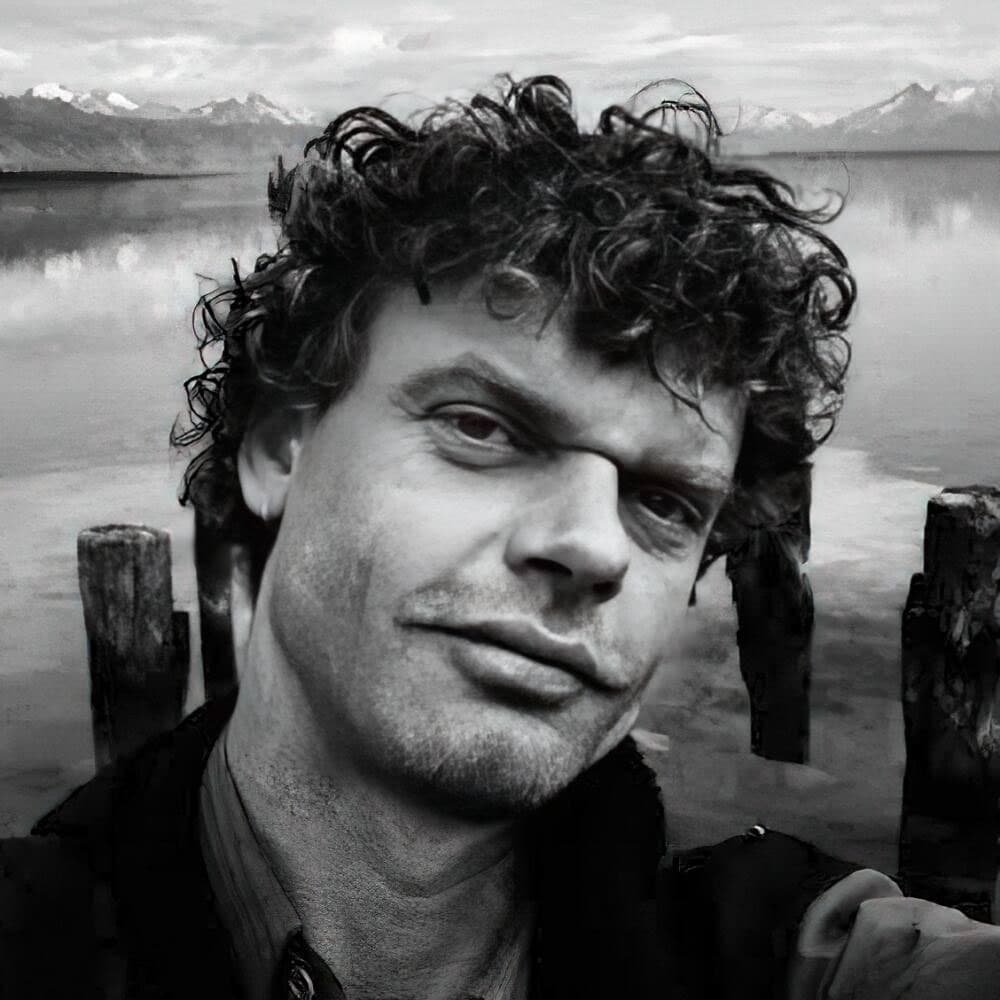 B&W self portrait of Kadir Van Lohuizen in front of a body of water with mountains in the distance