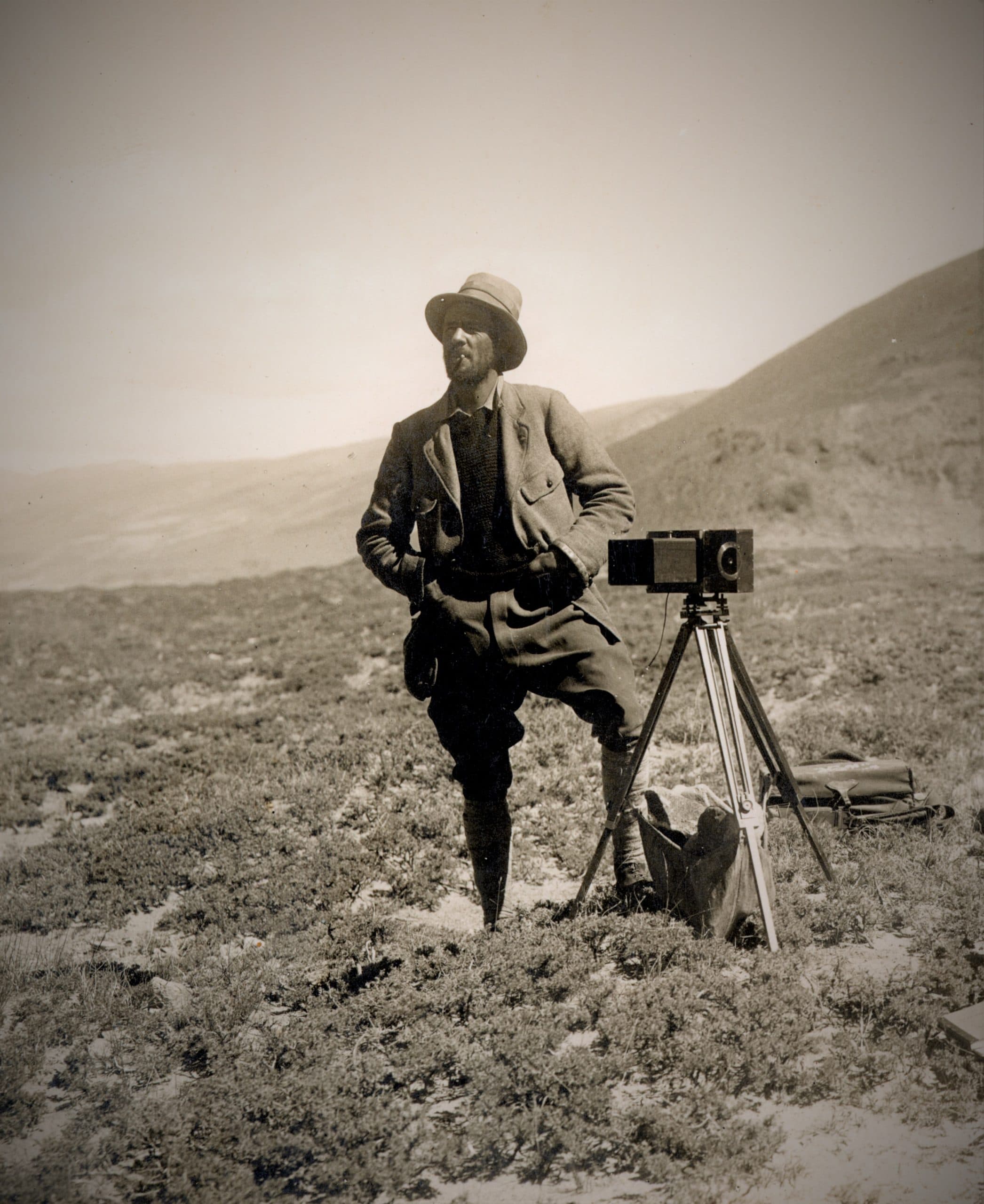 Sepia toned full-length portrait of Major E.O. Wheeler wearing a hat, jacket, pants, and high boots next to a camera on a tripod in a natural environment
