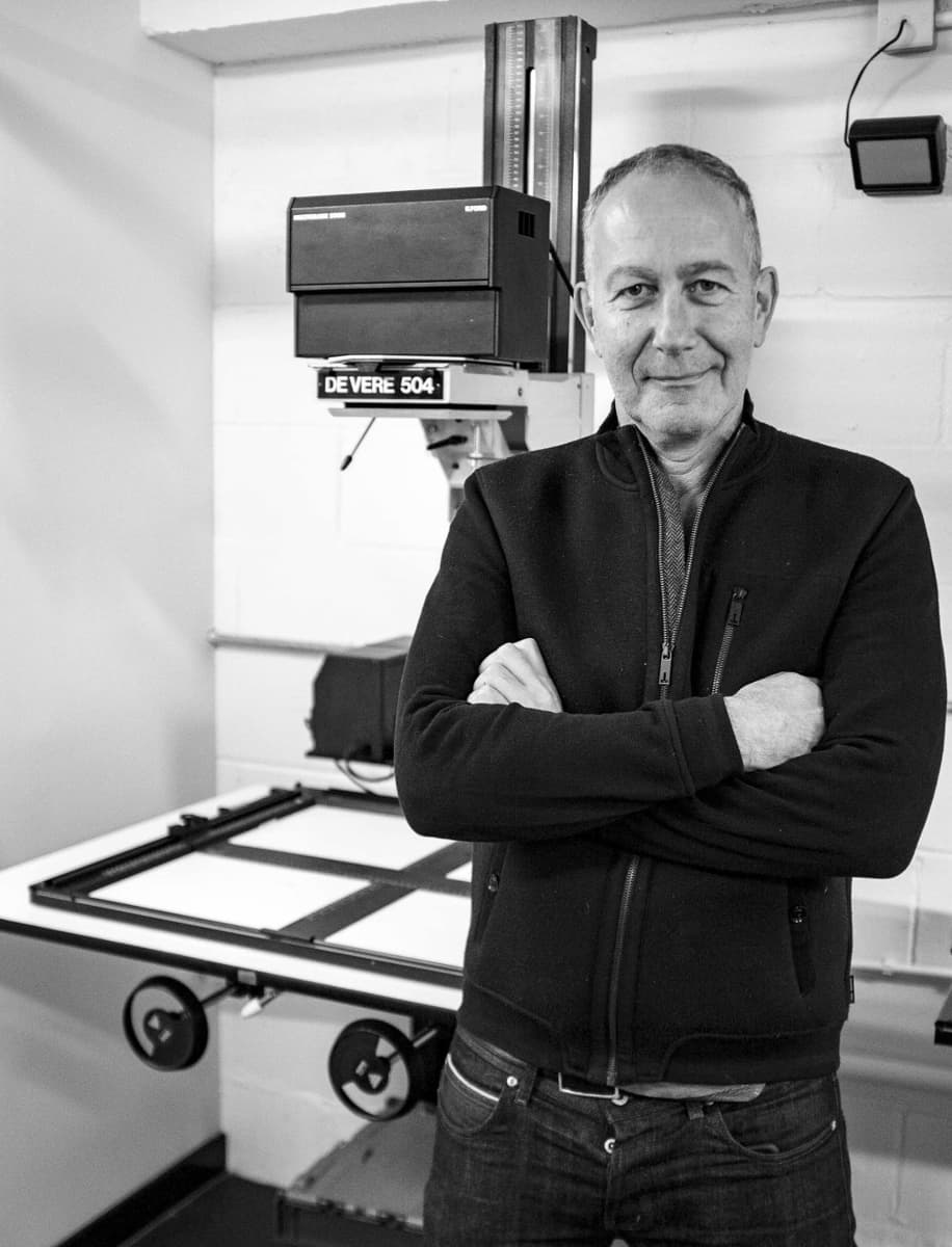 B&W portrait of Nadav Kander wearing a dark zipped jacket in front of a photographic enlarger