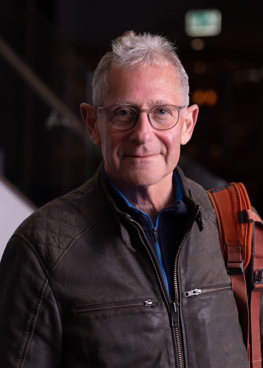 Color quarter length portrait of Stuart Franklin wearing grey glasses, a brown zipped leather jacket, and carrying an orange backpack on his left shoulder