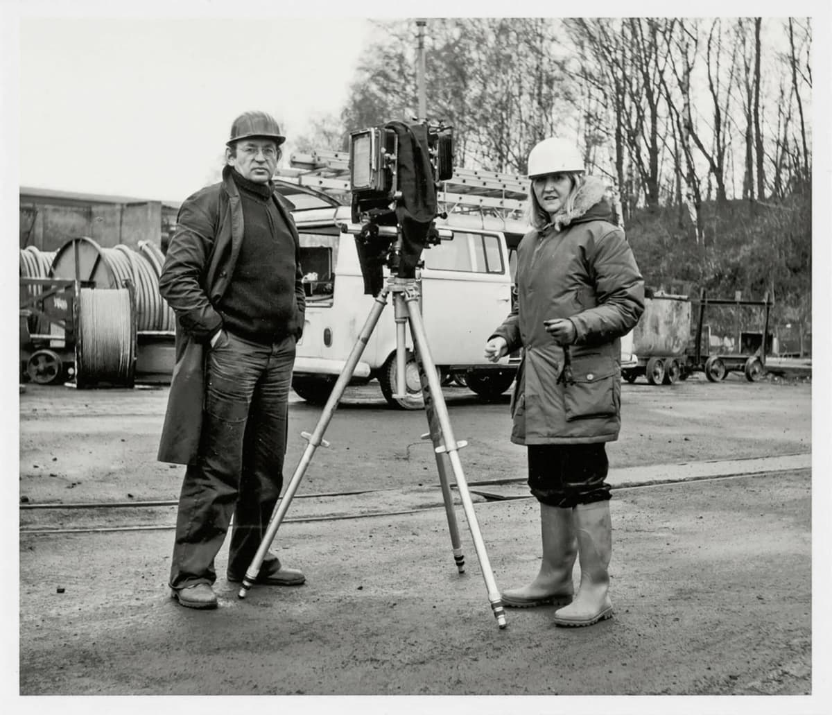B&W full length photo of Bernd and Hilla Becher next to a large format camera on a tripod, both wearing coats and hard hats